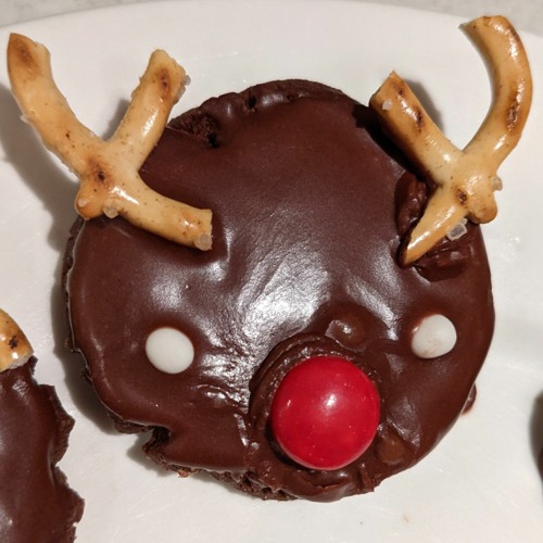 A decorated Rudolf the Reindeer biscuit with brown chocolate icing, a red m&m nose, two little white icing eyes and parts of pretzles for antlers